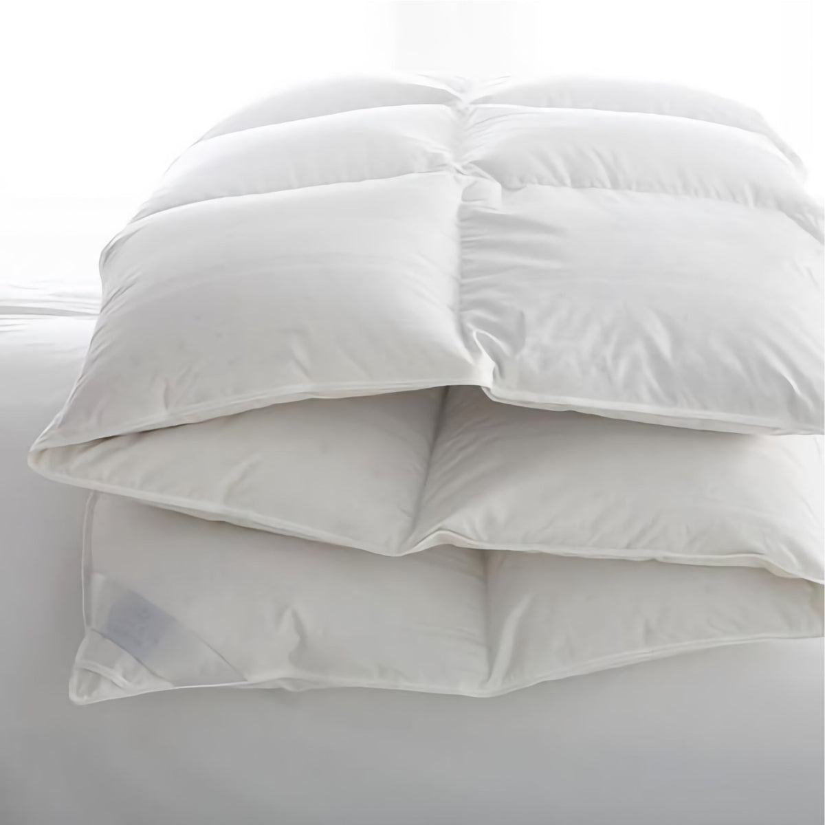 A white, fluffy comforter is folded neatly in thirds on a white bed, with soft natural light coming from the background.