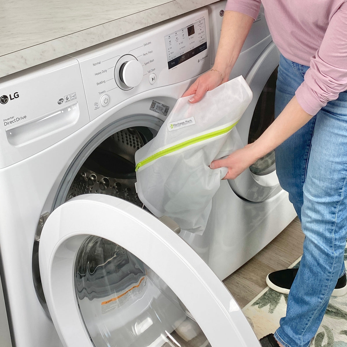 A person wearing jeans and a pink shirt puts Heritage Park Premium Fine Mesh Laundry Bags into a front-loading washing machine in a modern laundry room.