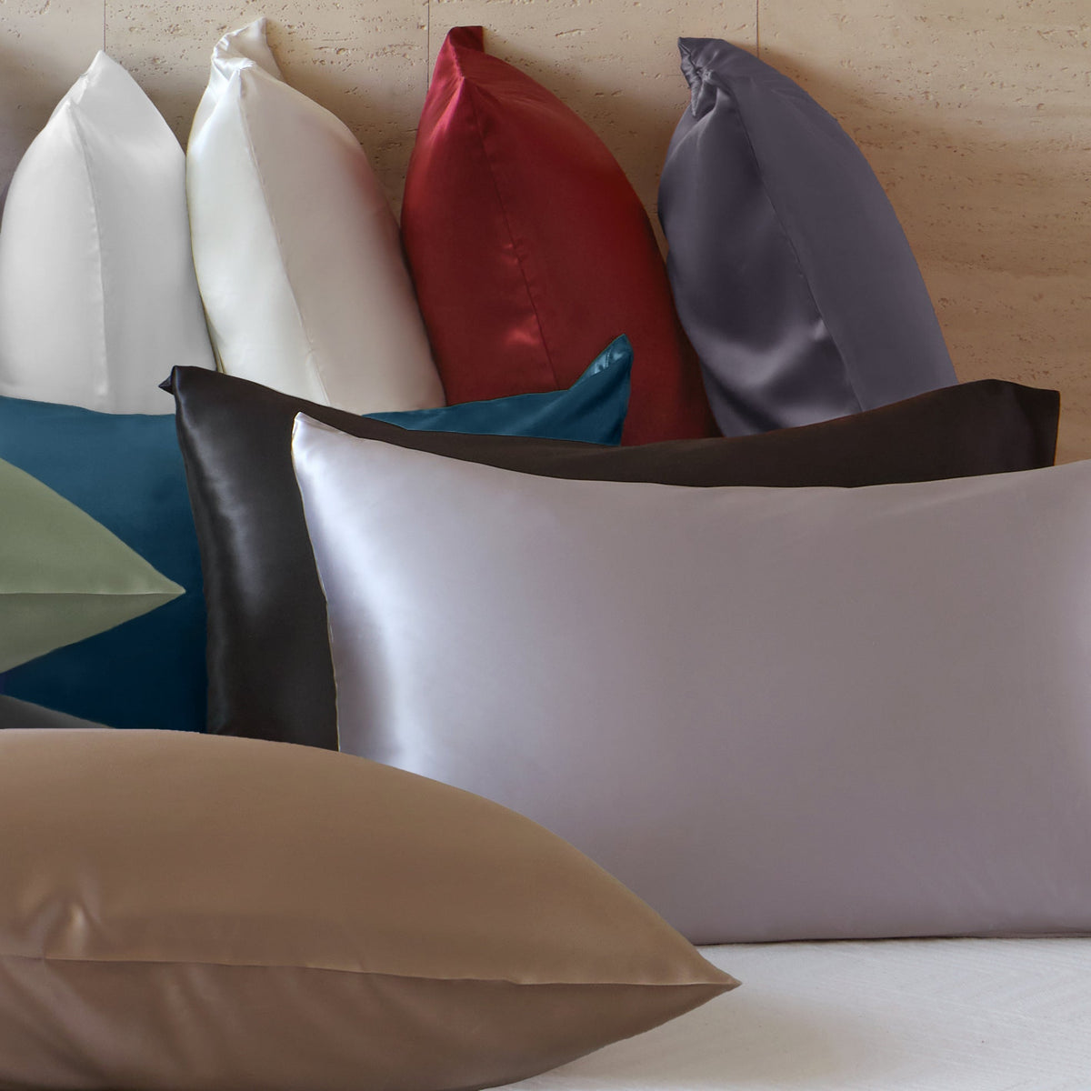 Several pillows with smooth, shiny pillowcases in various colors, including white, black, gray, red, purple, teal, brown, and beige, are arranged on a bed against a neutral wall.