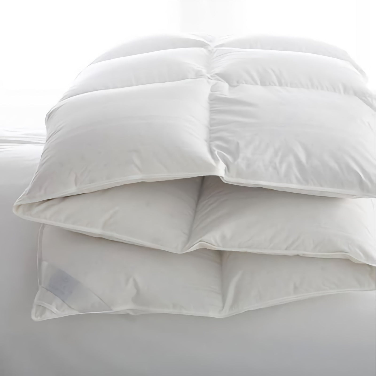 A white, fluffy comforter is folded neatly in thirds on a white bed, with soft natural light coming from the background.
