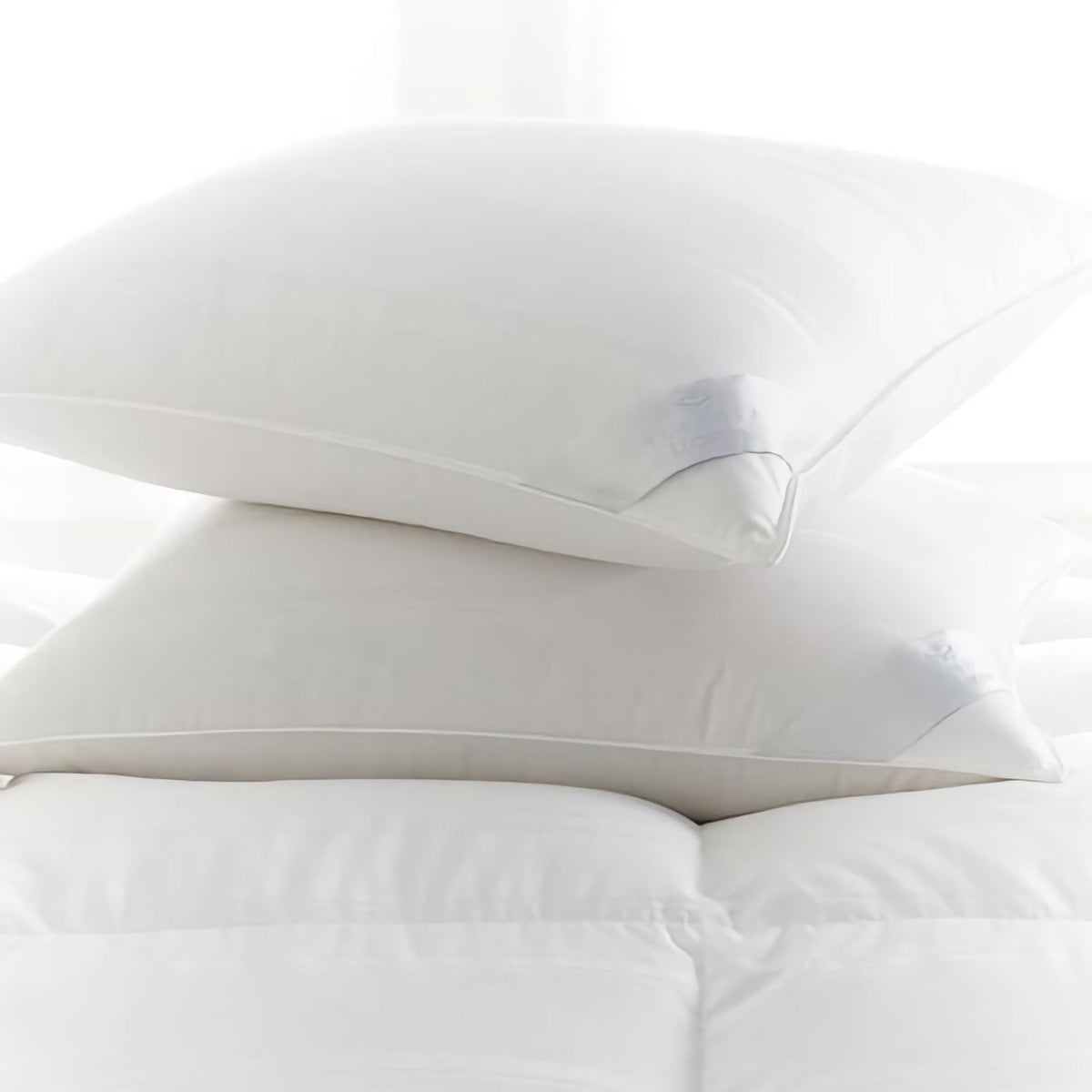 Two white pillows are stacked on top of each other on a white bed with soft, quilted bedding. The background is bright and softly lit, suggesting natural daylight.