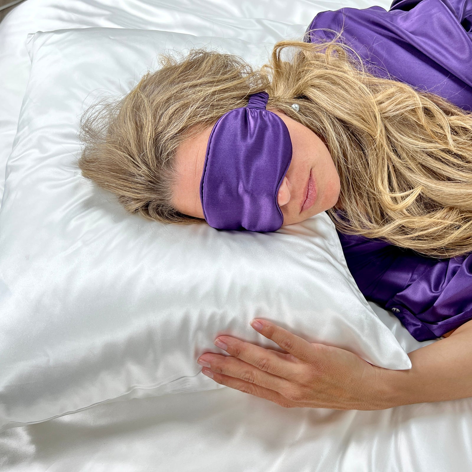 A person with long blonde hair lies on a white satin pillow, wearing a purple eye mask and purple satin sleepwear. Their hand rests on the pillow, and they appear relaxed.