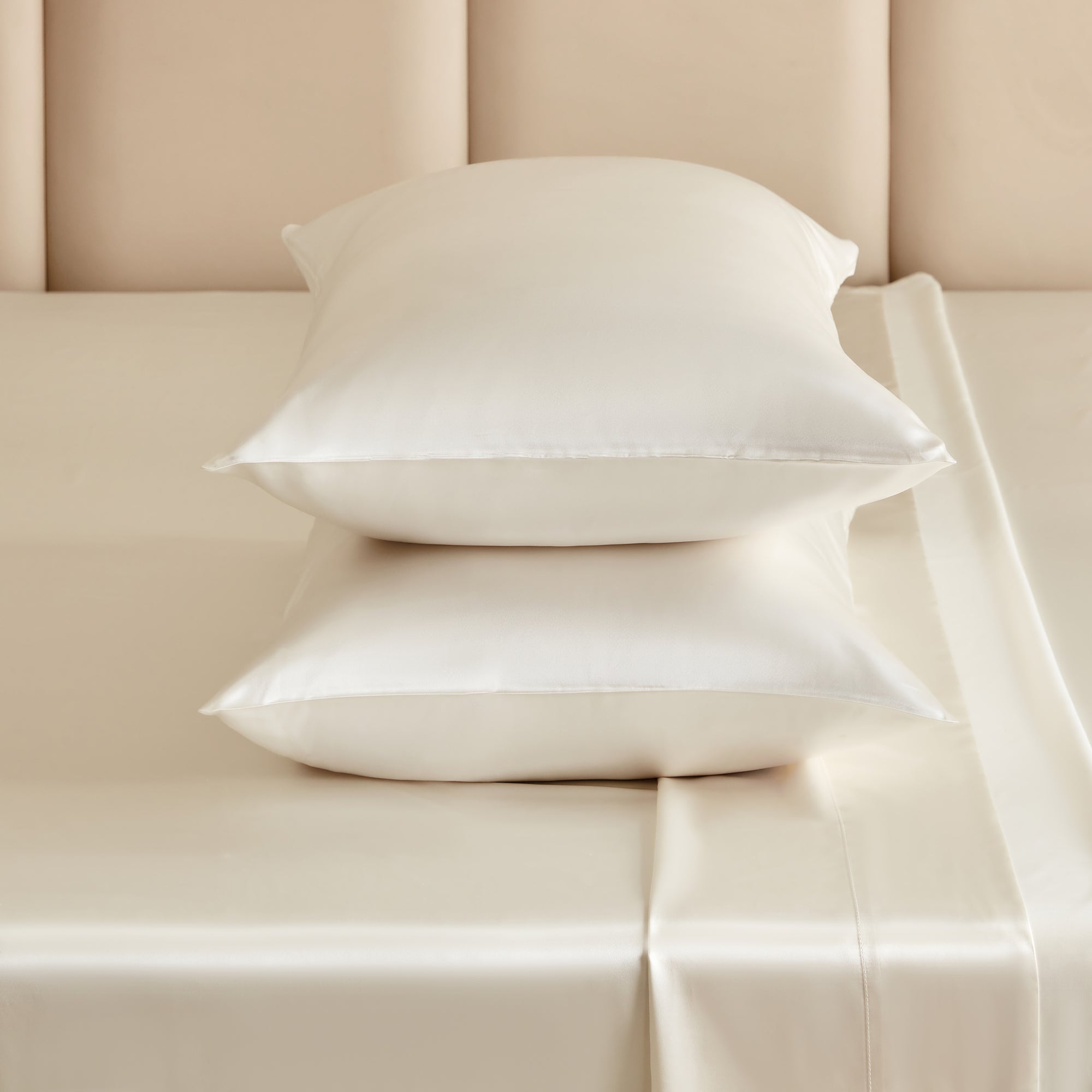 Two white silk pillows stacked on a matching sheet bed with a padded beige headboard in the background.