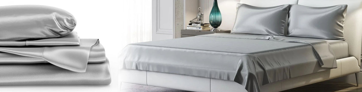 A neatly made bed with silver silk sheets and pillowcases next to a stack of folded matching bedding.