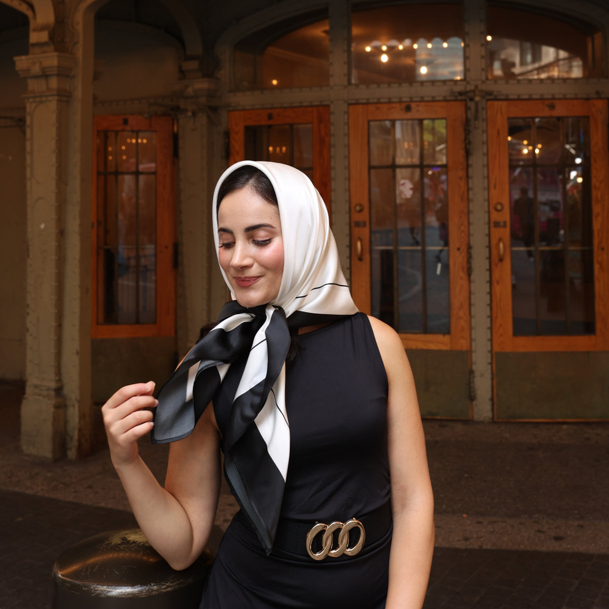 Young woman outdoors wearing a Silk Square Head Scarf in Black/Ivory from Mulberry Park Silks, styled elegantly with a sleeveless dress and gold-ring belt.