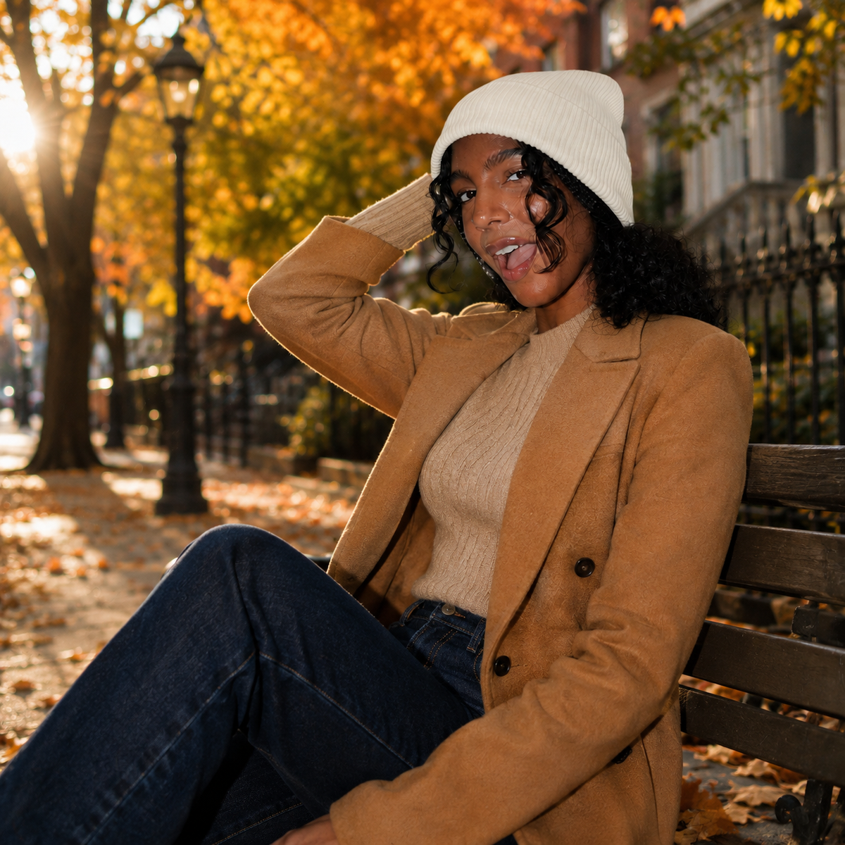 Woman in tan coat and wool cashmere beanie sits on a bench in a park with autumn leaves and sunlight behind her.