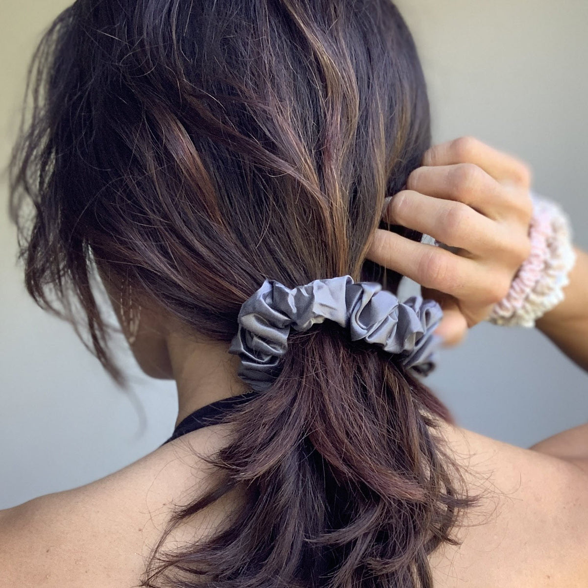 A person with long brown hair gathers it with a gray silk scrunchie, holding it with their right hand.