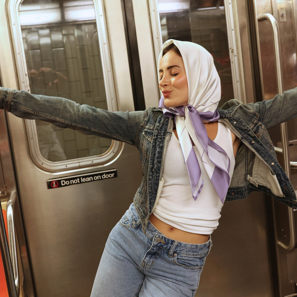 Young woman wearing Mulberry Park Silks' Silk Square Head Scarf in Lilac/Ivory, stylishly tied over her head, inside a subway car.