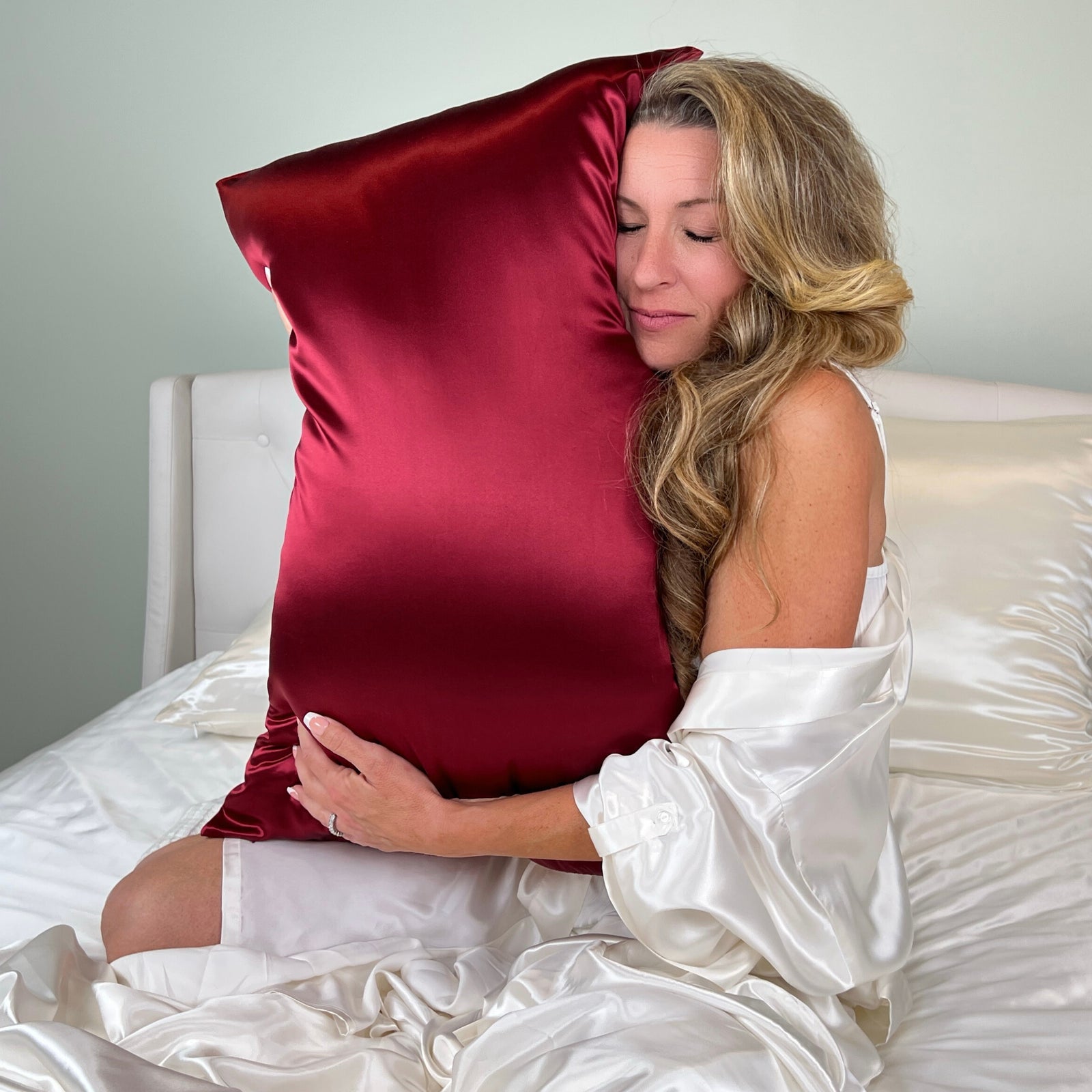 Woman sitting on a bed dressed in a ivory robe, hugging a rich Cabernet red Mulberry Park Silks pillowcase made from 100% pure Mulberry silk, showcasing a soft sheen and luxurious texture.
