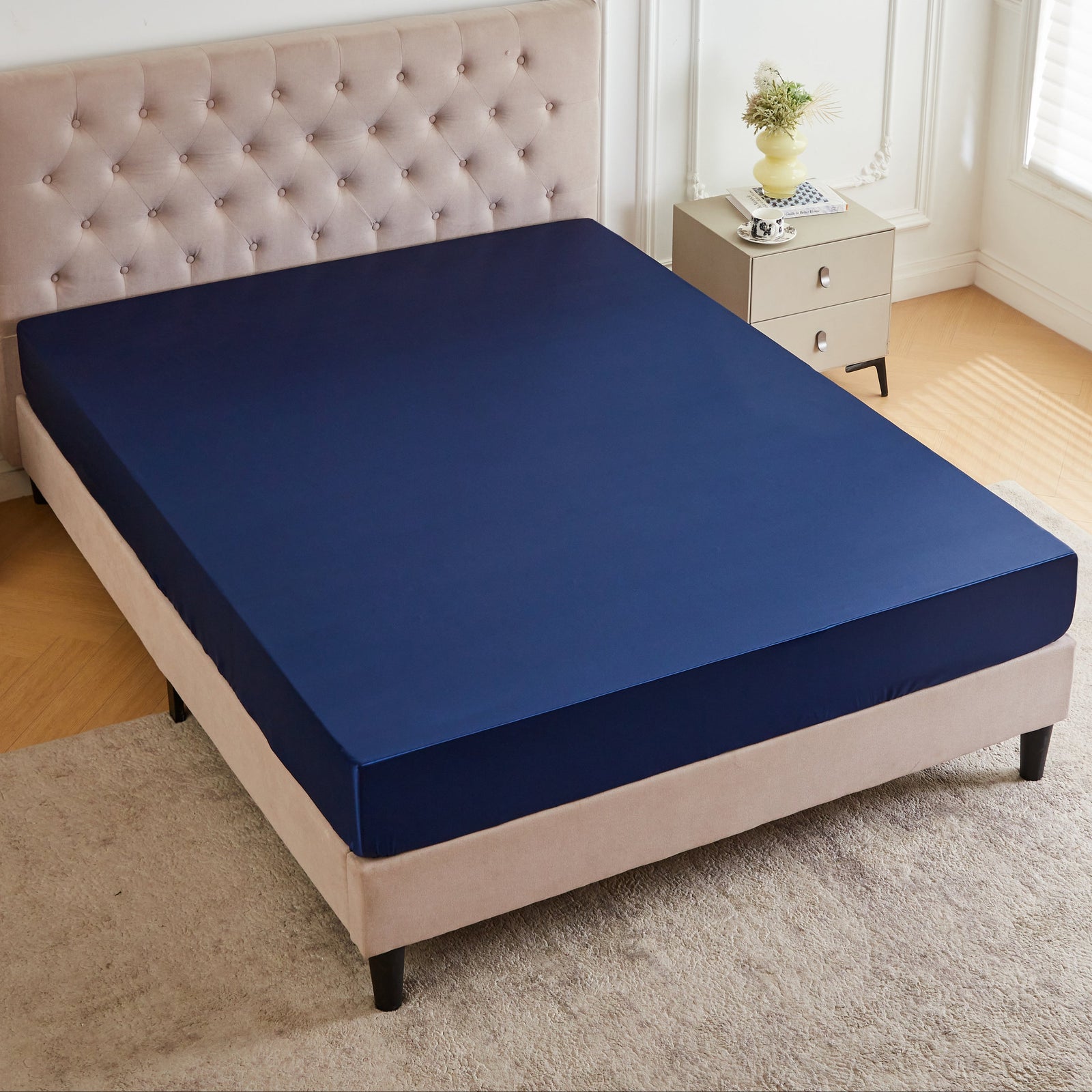 A neatly made bed with a 30 Momme Silk Fitted Sheet in navy, beige tufted headboard, and a vase on the nightstand.