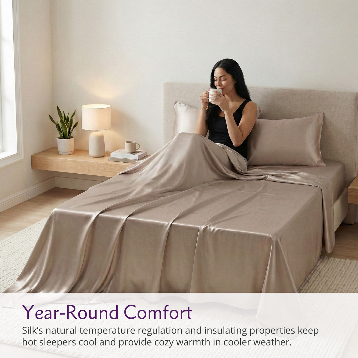 A woman sits on a bed with 22 momme silk sheets, holding a mug, in a bright, minimalist bedroom.