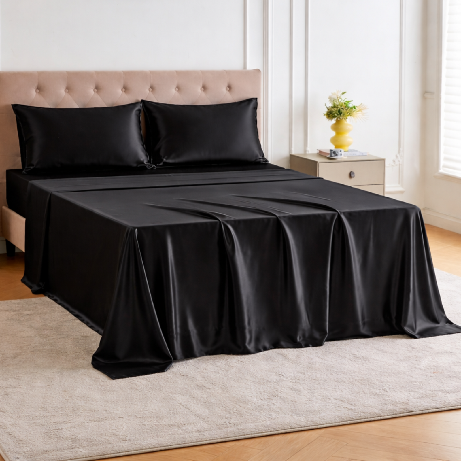 A bed with luxury silk bedding and black satin pillows, beside a nightstand with a yellow vase and flowers in a bright room.