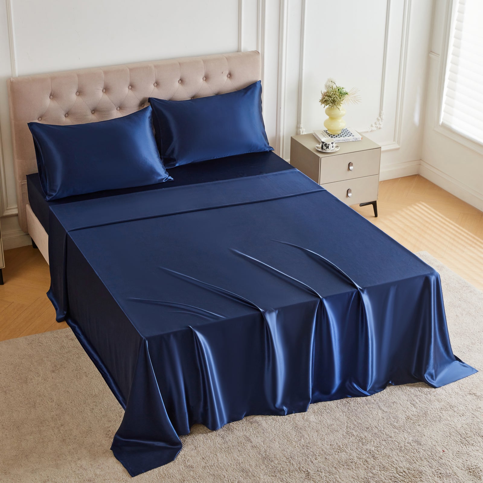 A bed with a navy blue silk sheet set, two pillows, and a nightstand with a vase in a bright bedroom.