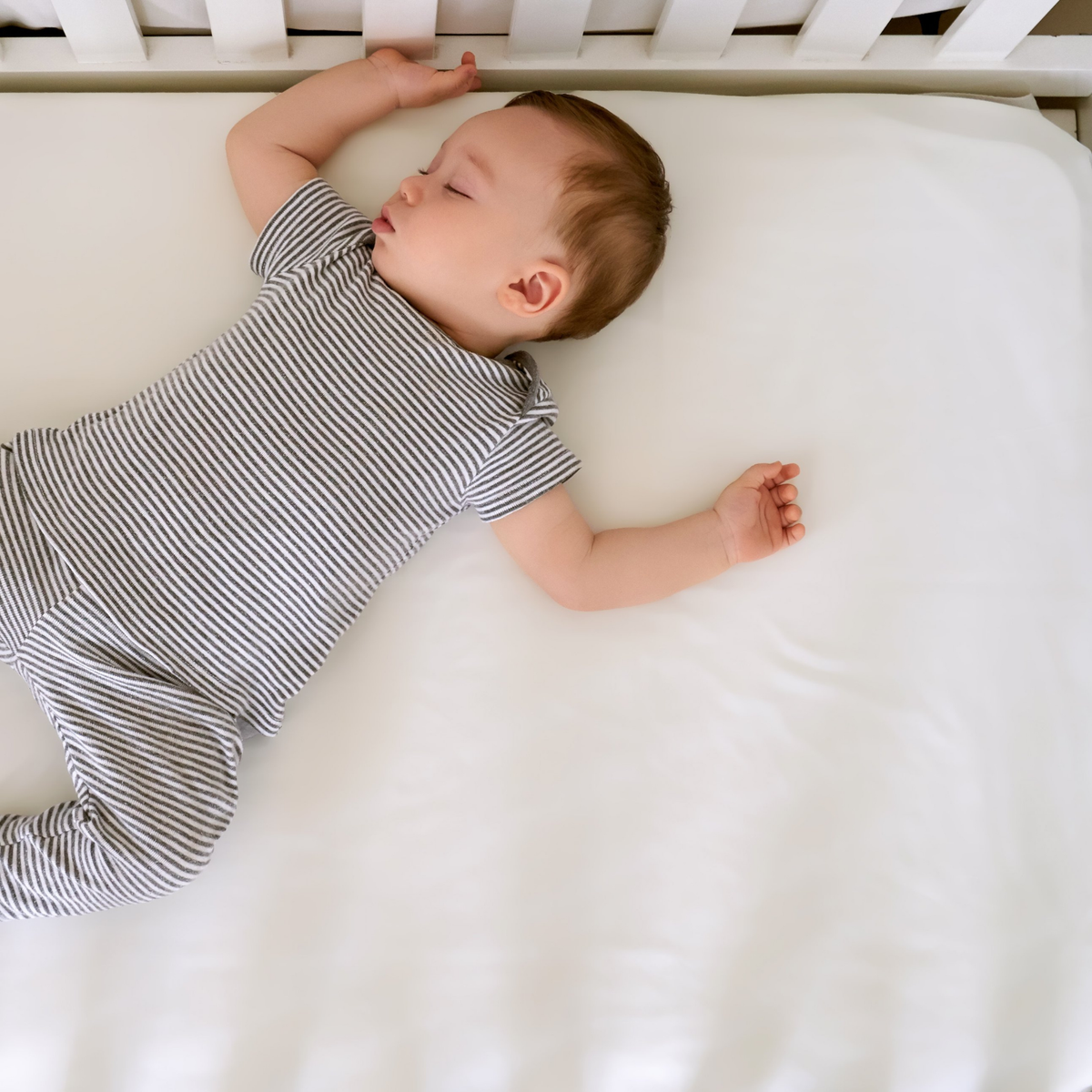 A baby sleeps on a white silk sheet in a crib, wearing a short-sleeved, gray and white striped onesie.