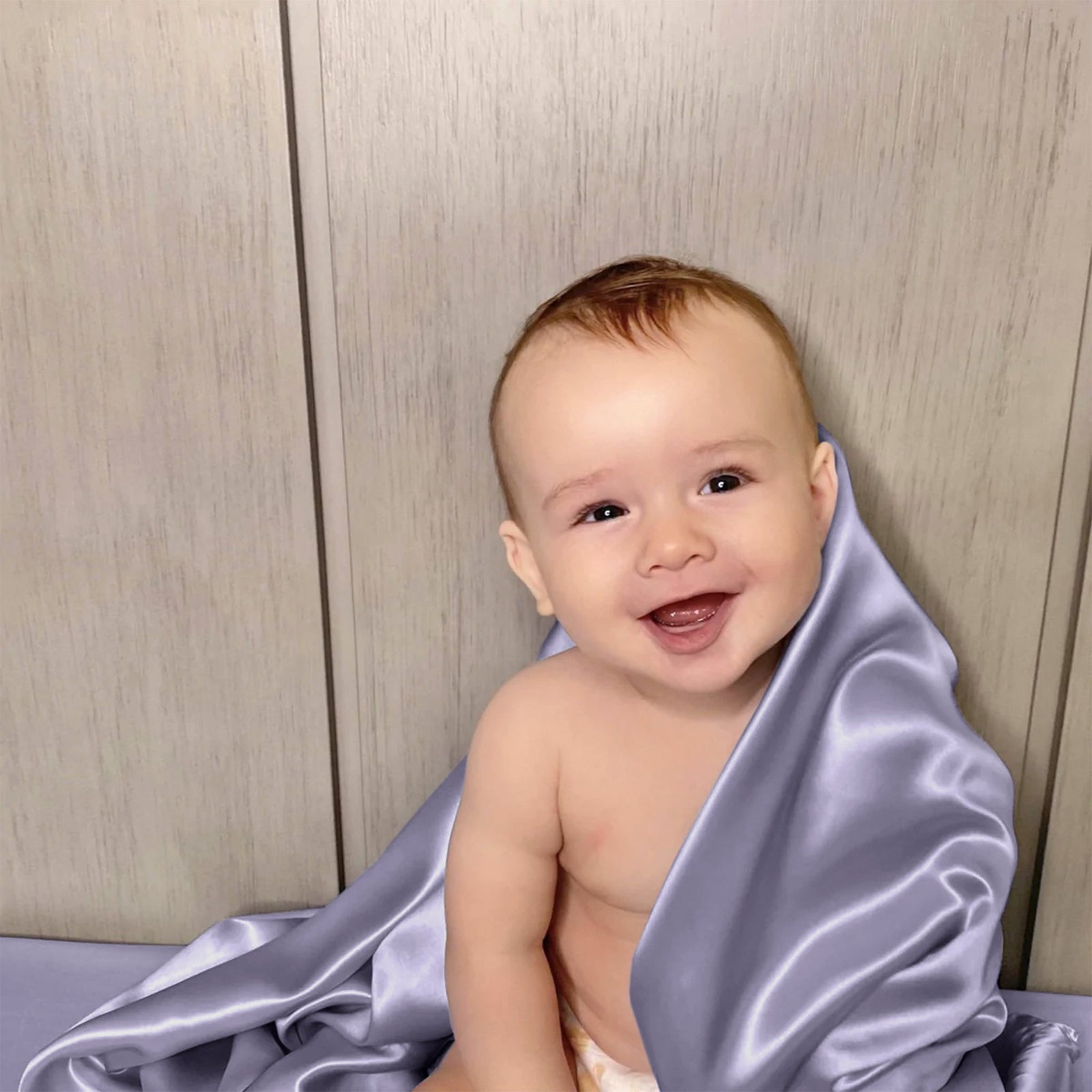 White crib with lavender light purple silk crib sheet on the mattress, a pillow, and doll in a room with a plant and framed picture.