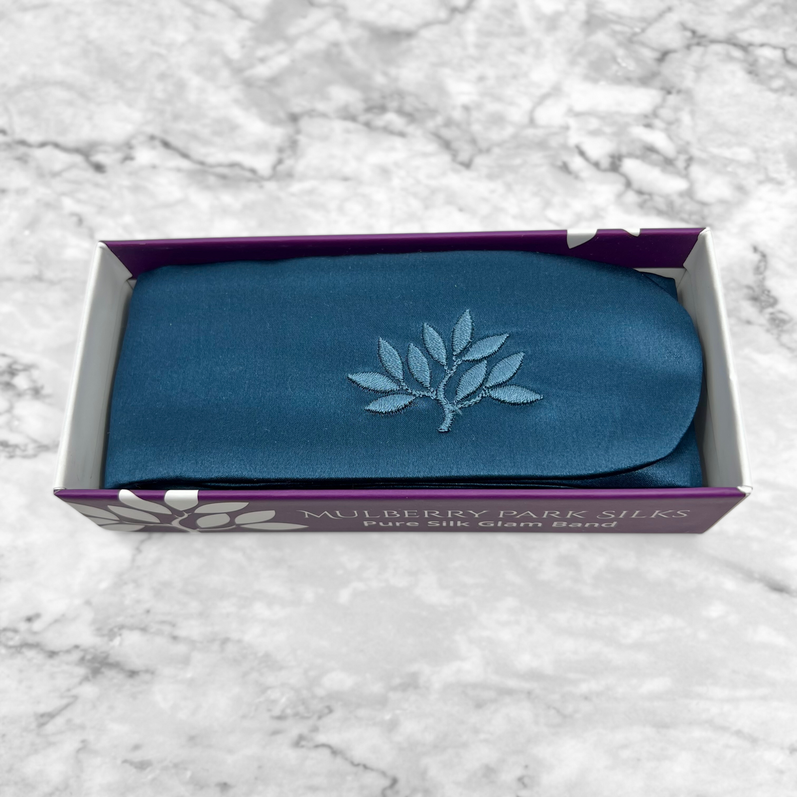 Mulberry Park Silks Pure Silk Glam Band in Deep Sea, folded in a purple-and-white box, featuring a leafy branch motif on a white marble surface.