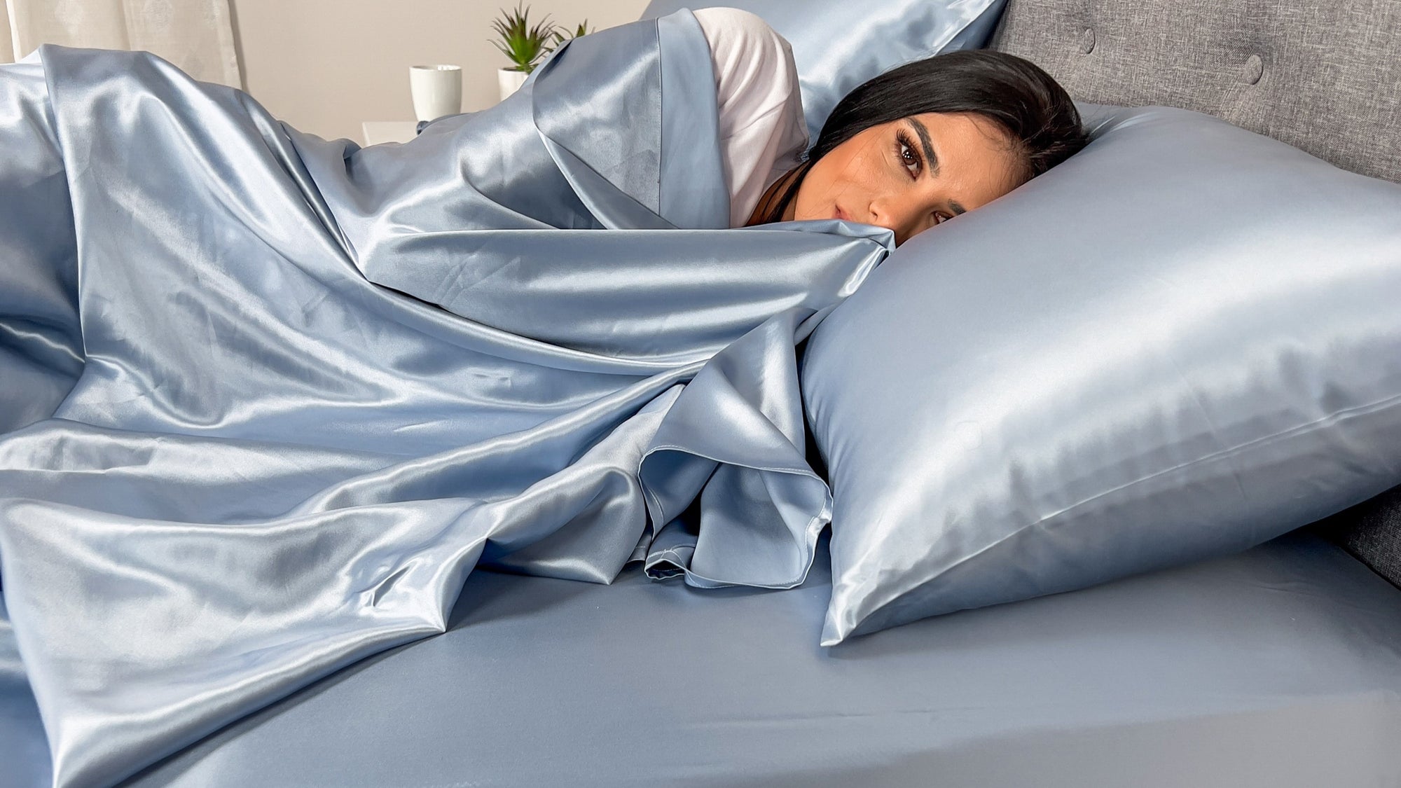 A woman lies in bed under light blue silk sheets, resting her head on a matching silk pillow.