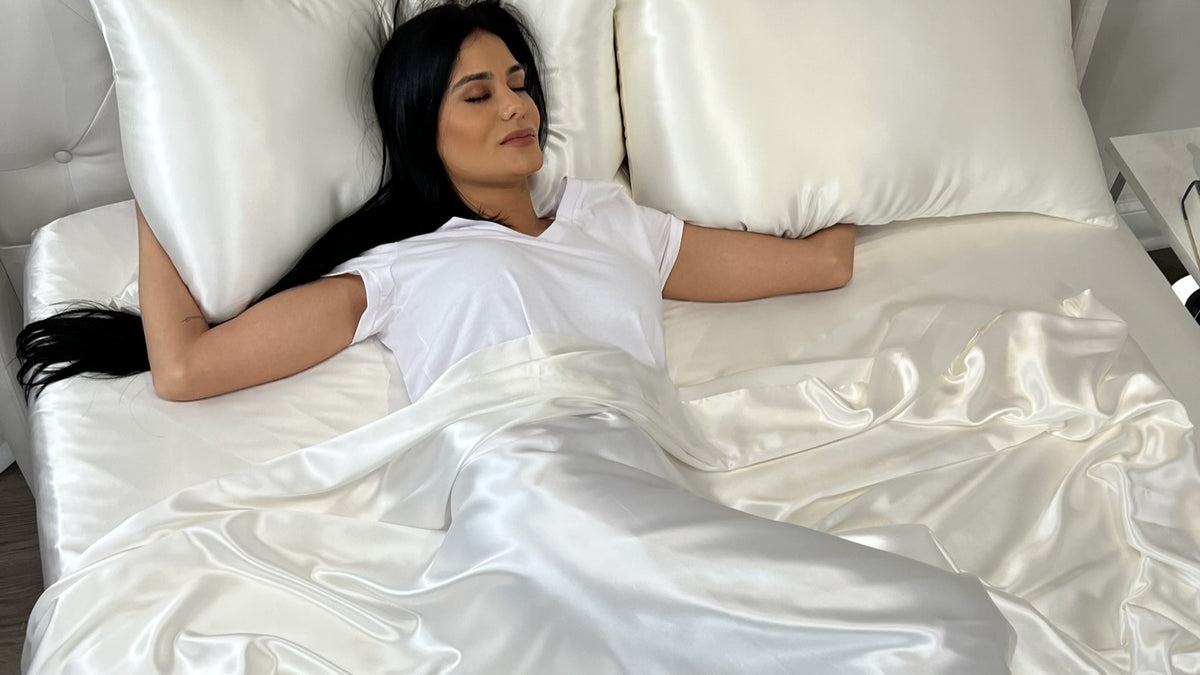 A woman with long dark hair sleeps on a bed with white silk sheets and pillows, wearing a white t-shirt.
