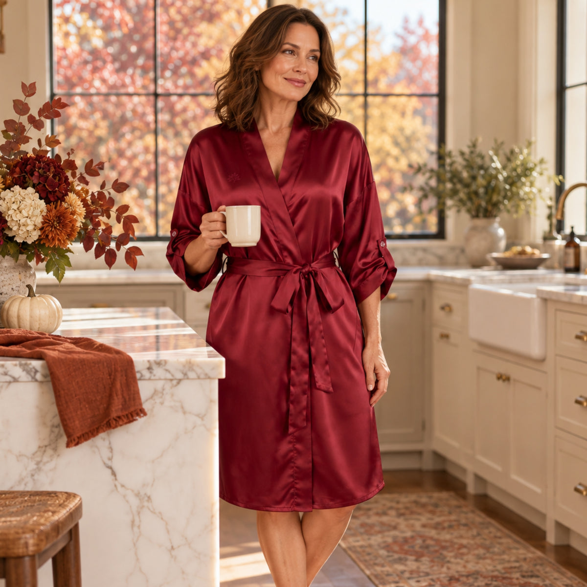 A woman in a burgundy 22 momme silk kimono robe holds a mug in a kitchen.
