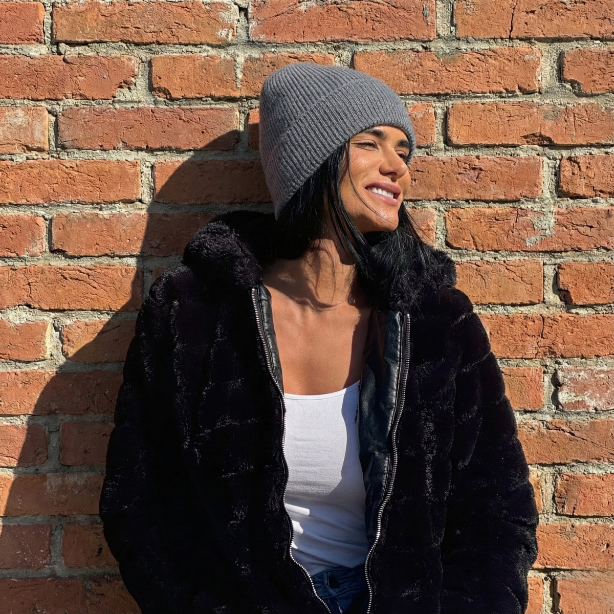 A woman wearing a black coat, gray beanie, and white shirt against a brick wall.