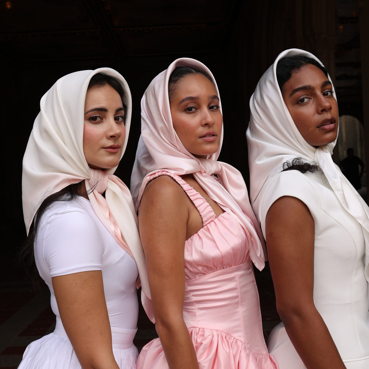 Three women wear Mulberry Park Silks' pink/ivory silk square head scarves, styled under the chin, showcasing elegance and versatile fashion.
