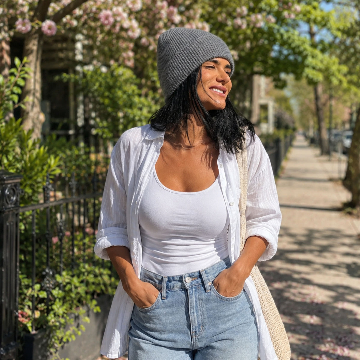 A woman in a silk lined wool cashmere beanie, white shirt, and jeans stands on a sunny, tree-lined sidewalk.