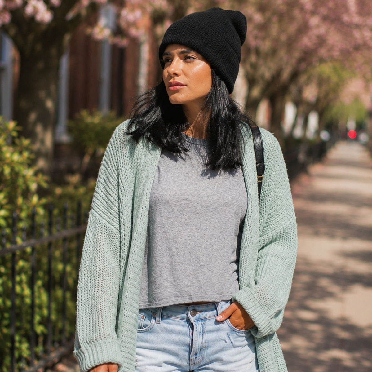 Woman wearing a black silk lined beanie, gray shirt, and light cardigan walking outdoors on a tree-lined sidewalk.
