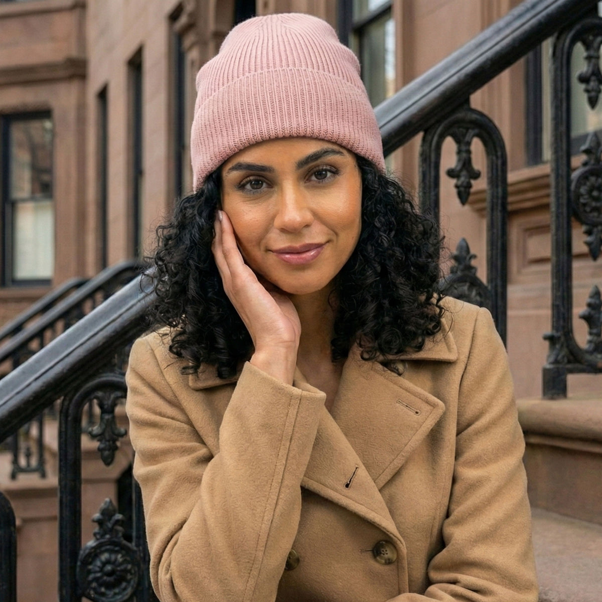 Woman in a tan coat and pink beanie sits on outdoor steps, hand on face, looking at the camera.