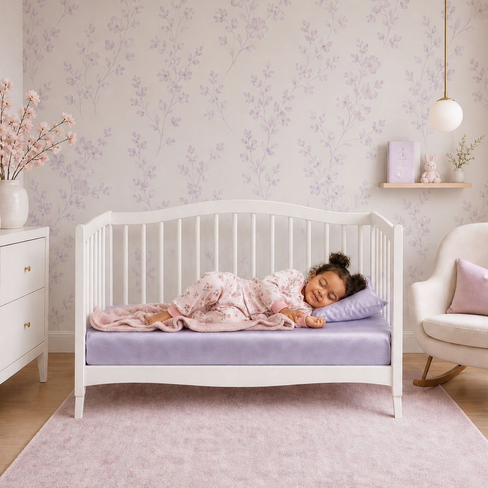 White crib with lavender light purple silk crib sheet on the mattress, a pillow, and doll in a room with a plant and framed picture.