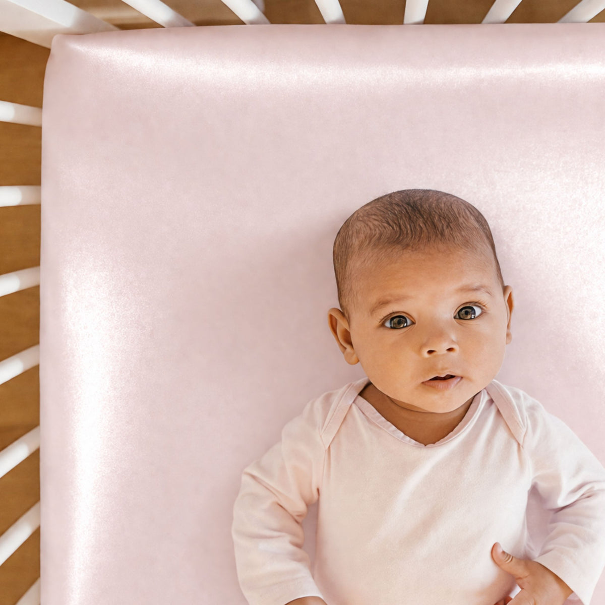 Baby in a light pink outfit lies on a Mulberry silk crib sheet inside a white crib, looking up at the camera.