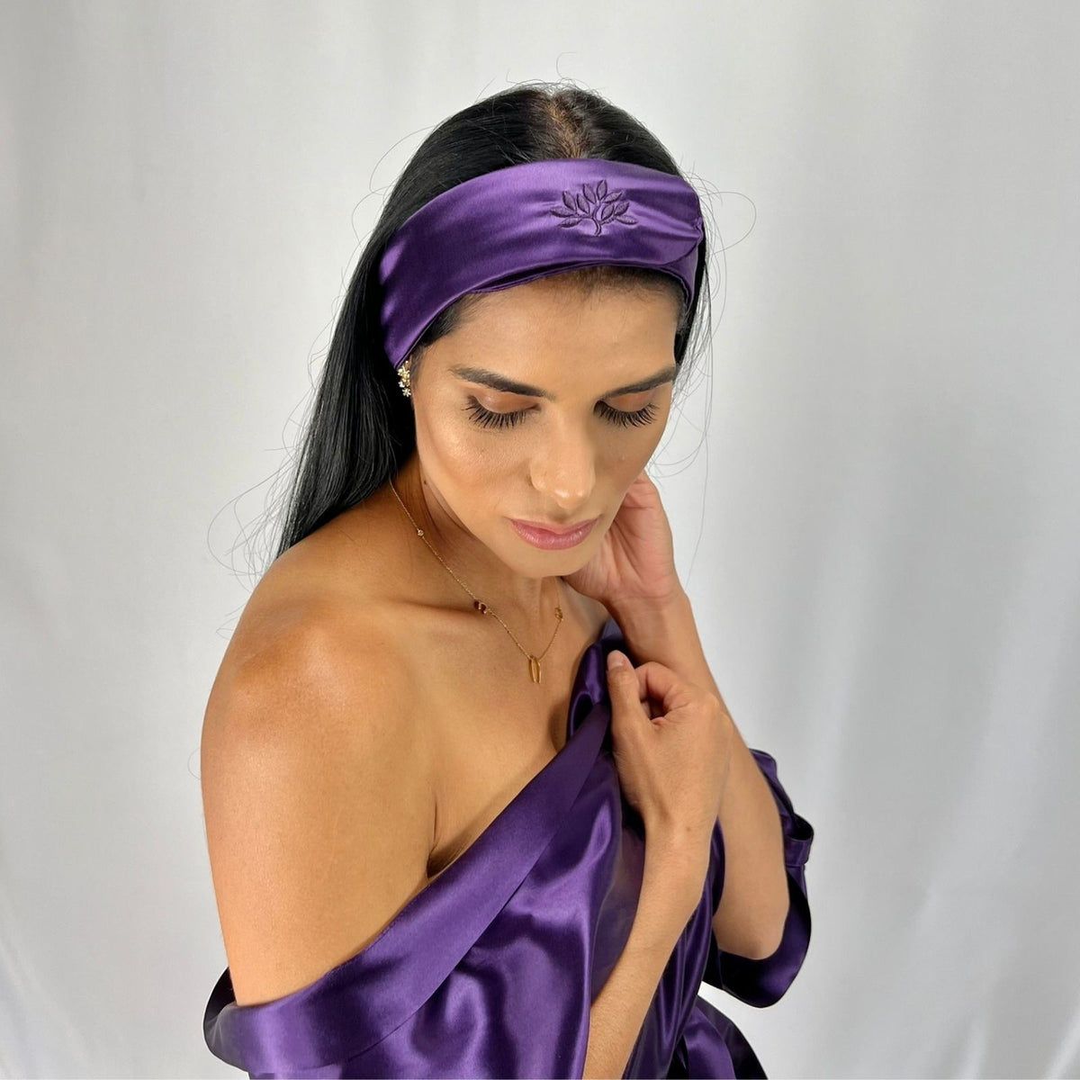 Woman with long dark hair wears a mulberry silk headband and matching off-shoulder dress, looking down.