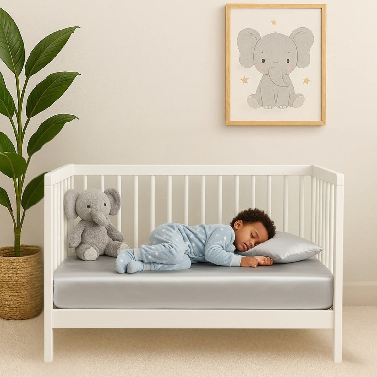 A toddler sleeps on a gray Mulberry silk crib sheet in a white crib with an elephant toy and matching wall art.