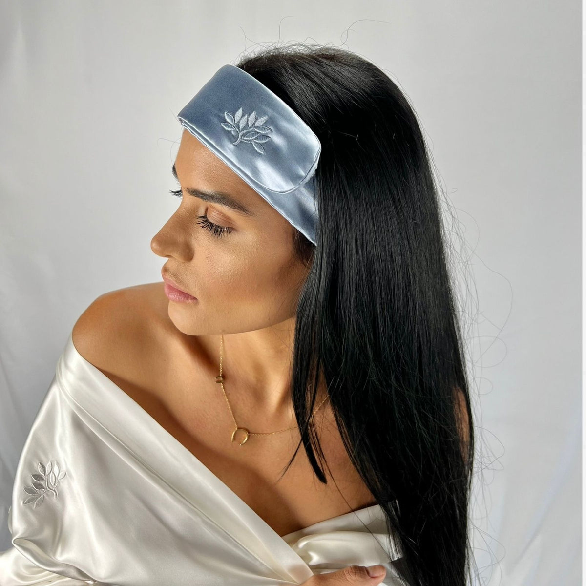 Woman with long dark hair wears a blue Mulberry silk headband and white off-the-shoulder top, looking to the side.
