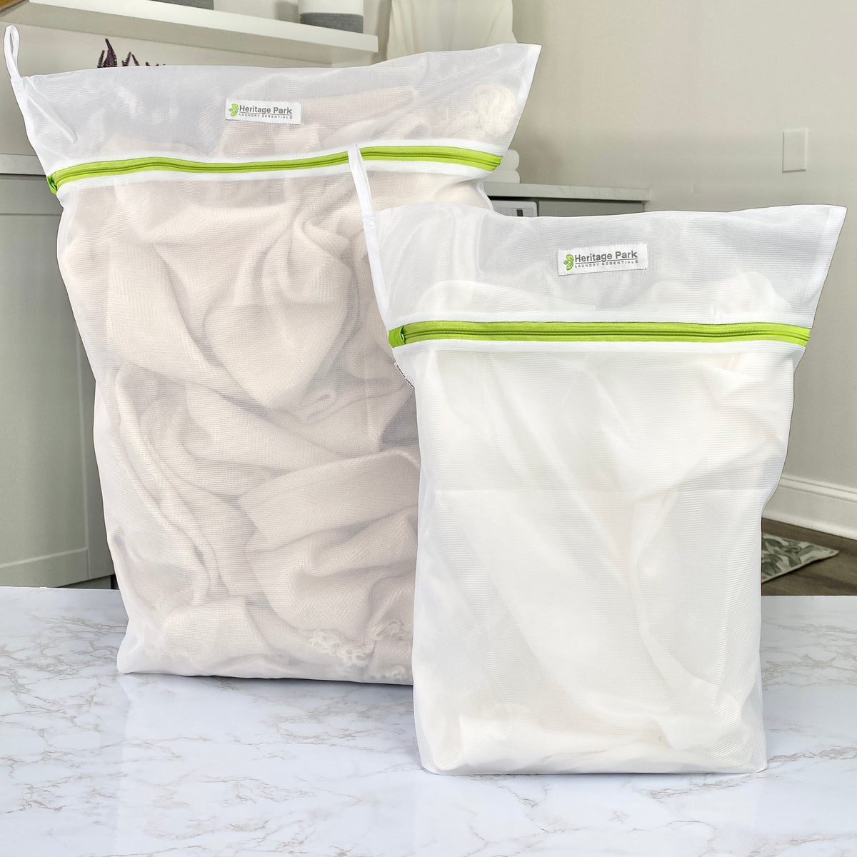 Two Heritage Park Premium Fine Mesh Laundry Bags, featuring green zippers and made from premium polyester mesh, are filled with light fabrics and placed on a marble countertop—ideal for protecting fine washables in any modern space.